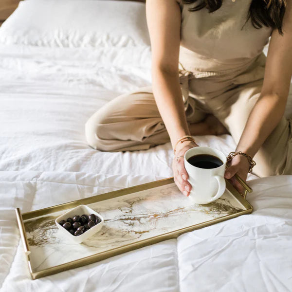 Person sitting on a bed holding a cup of coffee with a resin tray of coffee beans.