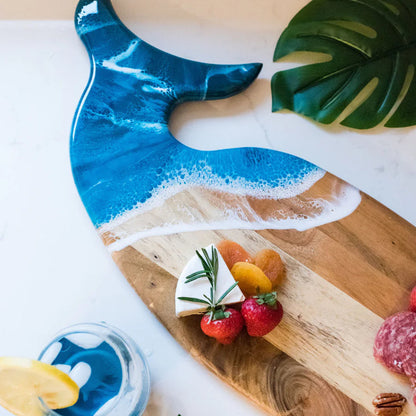 Wooden charcuterie board with blue resin design, surrounded by fruits and a glass of water on a light wooden surface.