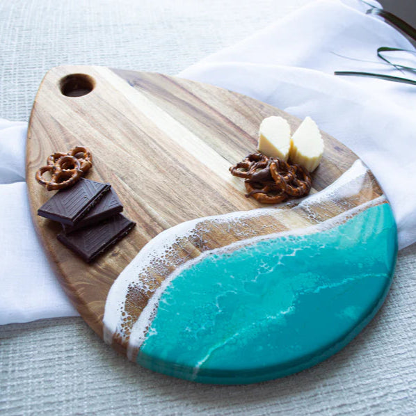 Wooden serving tray with a turquoise resin design, chocolate, and pretzels on a white cloth.