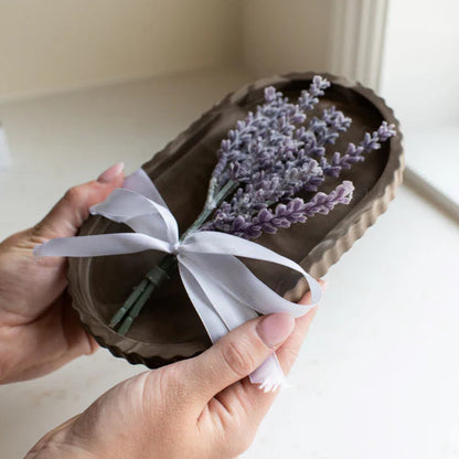 Brown sandal with lavender flowers and a white ribbon held by a hand on a light surface.