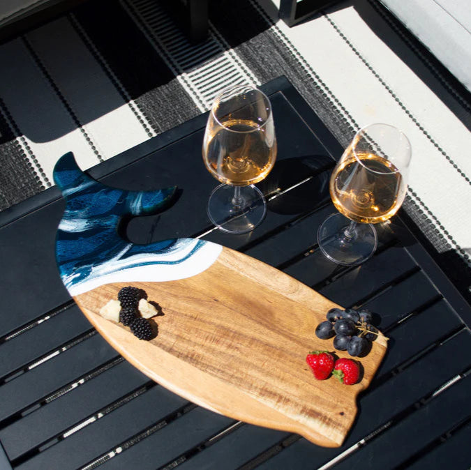 Two glasses of white wine on a wooden whale shaped charcuterie board with fruits and blackberries on a black surface.