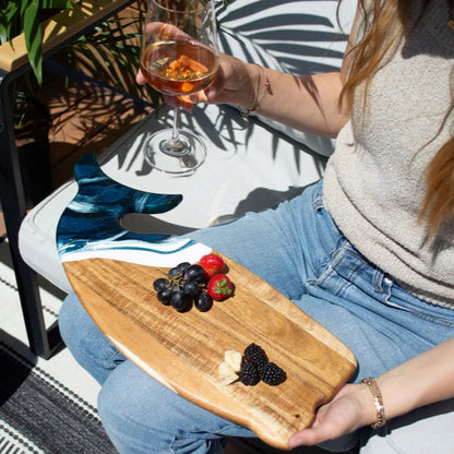 Person sitting outdoors with a wooden whale shaped charcuterie board holding fruits and a glass of wine.