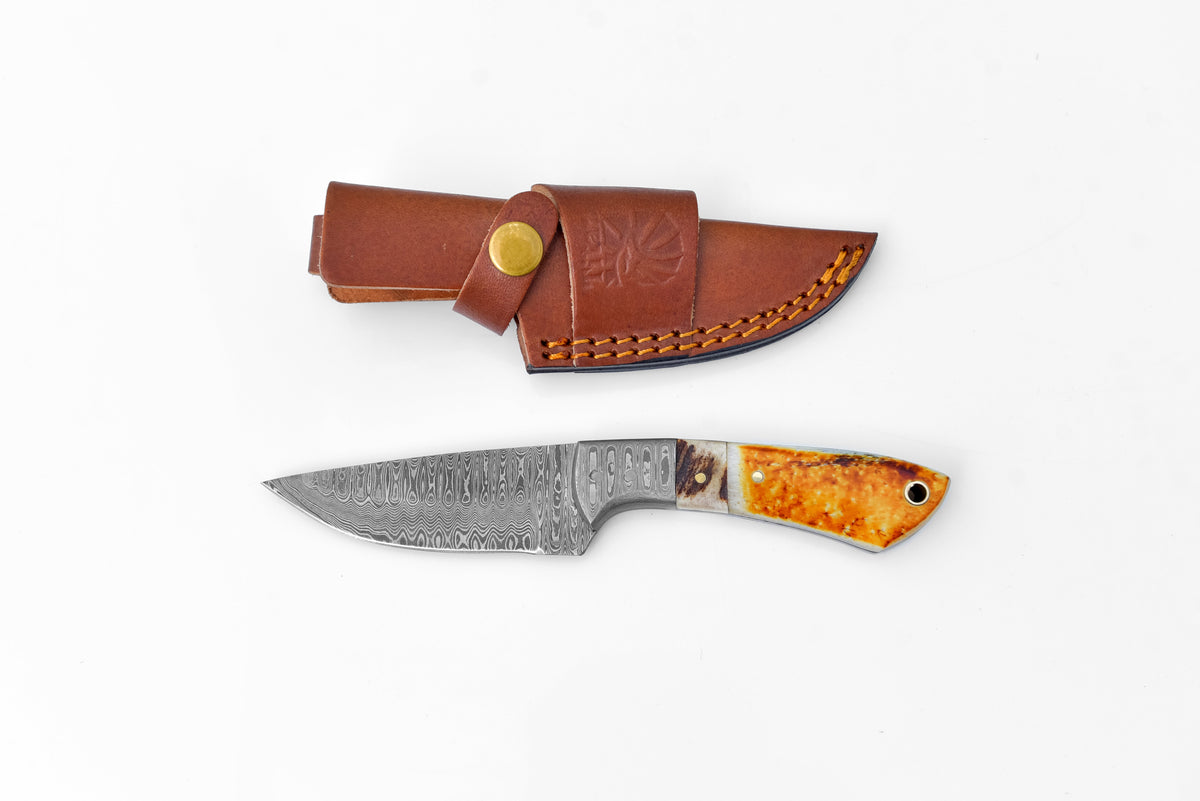 Titan Scorched Timber Damascus EDC Knife with burnt bone and stag horn handle resting alongside leather sheath