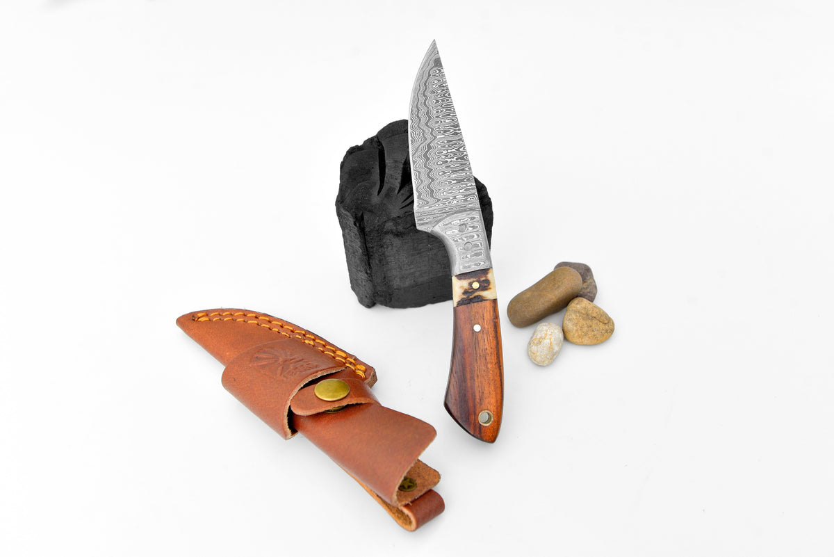 Damascus steel blade with intricate patterns and a smooth wooden handle, standing upright next to a brown leather sheath and small stones on white background.