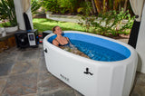 Woman relaxing in a white inflatable bathtub filled with clear water, set on a tiled patio near greenery and a black control unit.