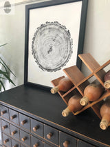 Black-framed print of a detailed black and white tree ring cross-section leaning against a wall, beside a wooden wine rack holding several bottles on a dark cabinet with multiple small drawers.