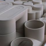 Matte beige ceramic double planter with smooth rounded edges and two cylindrical pots connected by a flat top surface, arranged among similar rounded ceramic vessels.