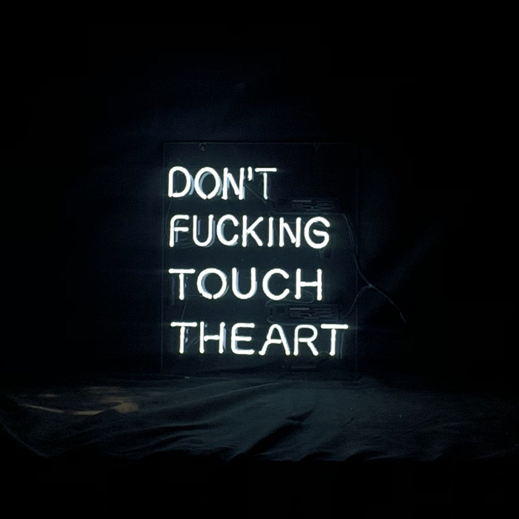 White neon sign with the phrase 'DON'T FUCKING TOUCH THE ART' in capital letters against a dark background, glowing softly on a black fabric surface.