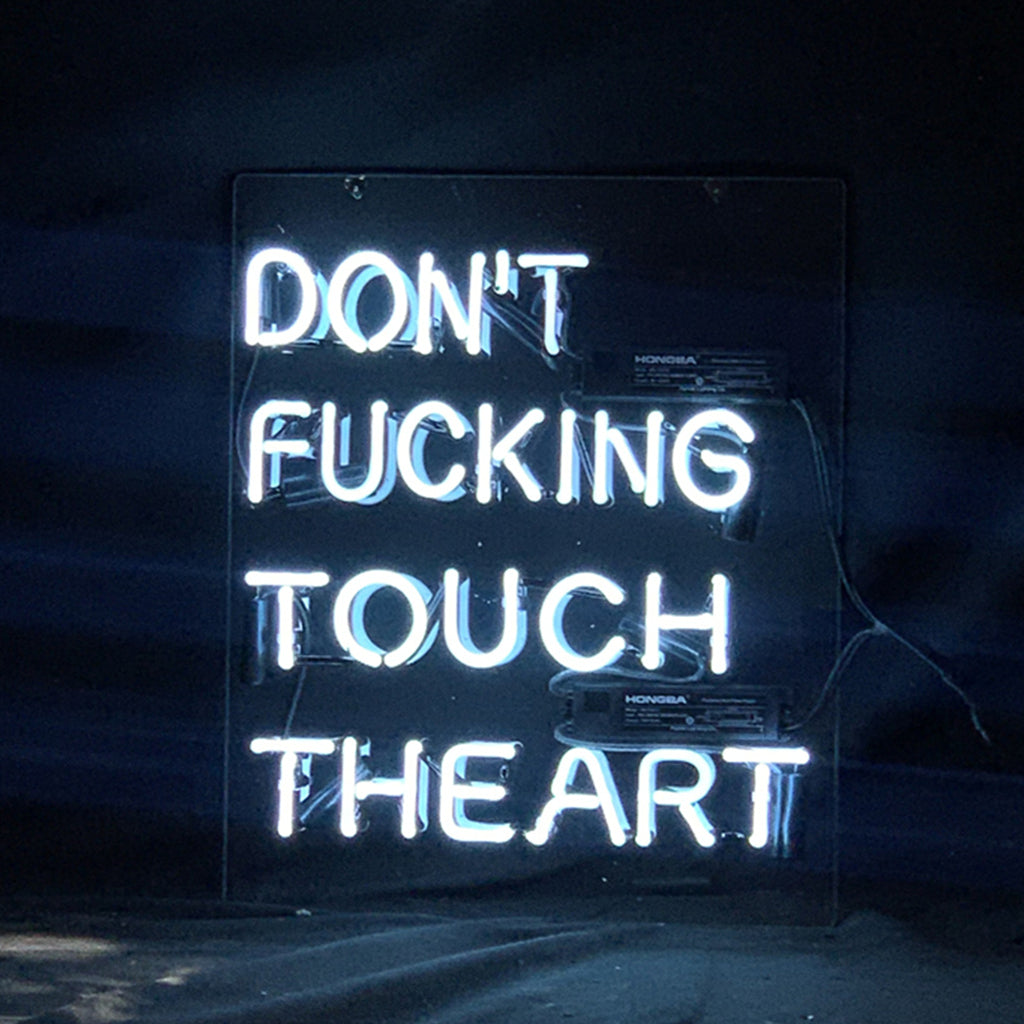 White neon sign on a clear backing reading 'DON'T FUCKING TOUCH THE ART' against a dark background with visible wiring and transformers