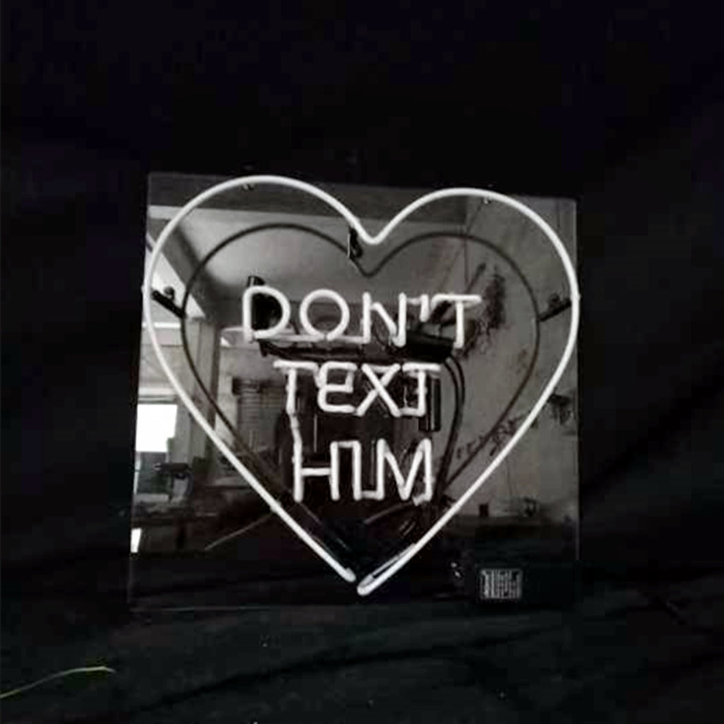 White neon tubing forming a heart shape around the text 'DON'T TEXT HIM' mounted on a reflective black square panel.