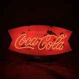 Red neon sign featuring the cursive Coca-Cola logo and the word 'DRINK' above it on a curved rectangular backing in a dark setting.