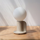 Minimalist Q Lamp with a frosted glass globe atop a matte beige curved base, casting soft shadows on a wooden surface under diffused natural light.