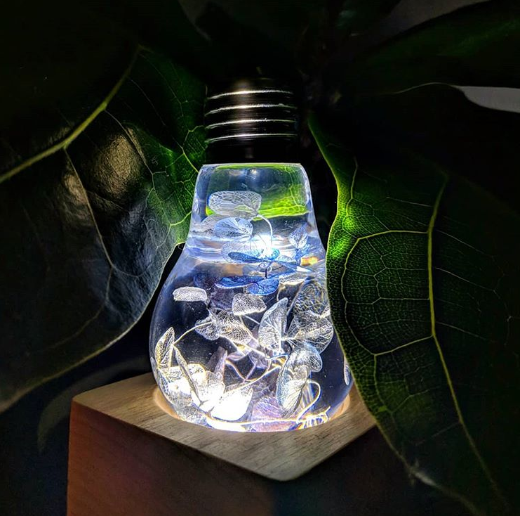 Light bulb-shaped lamp with engraved design on a wooden base, surrounded by leaves.
