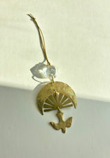 Gold-toned hanging ornament with hammered crescent and sunray design, topped by a clear vintage glass crystal and a dangling bird silhouette, casting shadows.