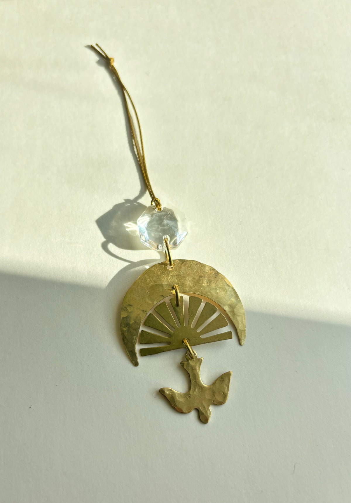 Gold-toned hanging ornament with hammered crescent and sunray design, topped by a clear vintage glass crystal and a dangling bird silhouette, casting shadows.