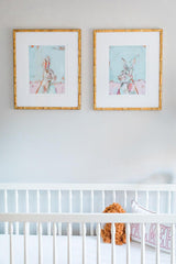Two framed Bunny Girl fine art prints by Chelsea McShane, each depicting a stylized rabbit with pastel colors and a light blue background, hung side by side above a white crib.