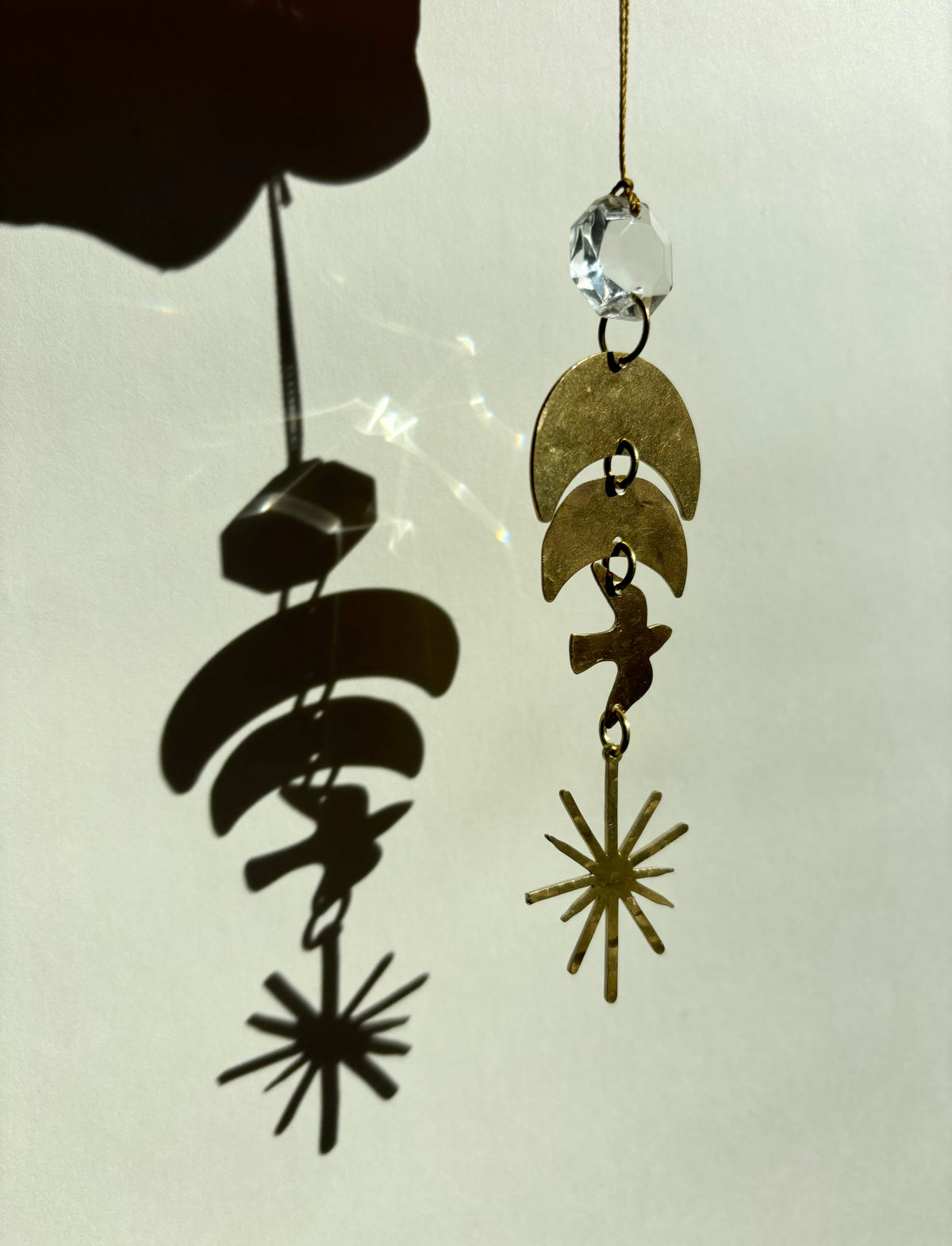 Brass pendant with crescent and star shapes hanging from a vintage glass crystal, casting a sharp shadow on a light background.