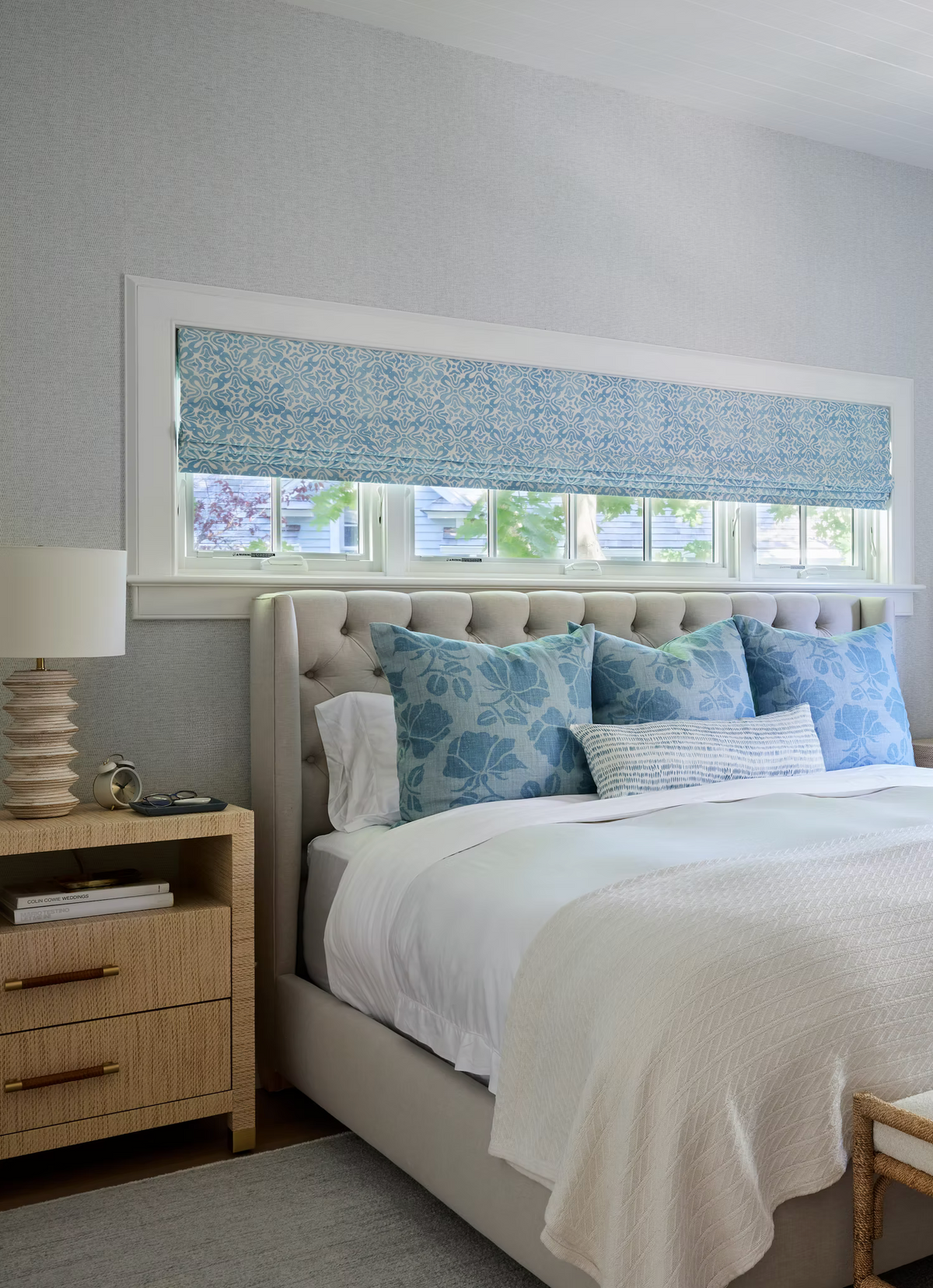 Bedroom featuring a tufted beige upholstered bed with blue floral pillows and a matching blue patterned Roman shade above a row of narrow windows.