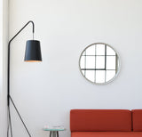 Round wall mirror with a thin beige frame reflecting a window with black grid, mounted above a rust-orange sofa and near a tall black floor lamp with a conical shade.