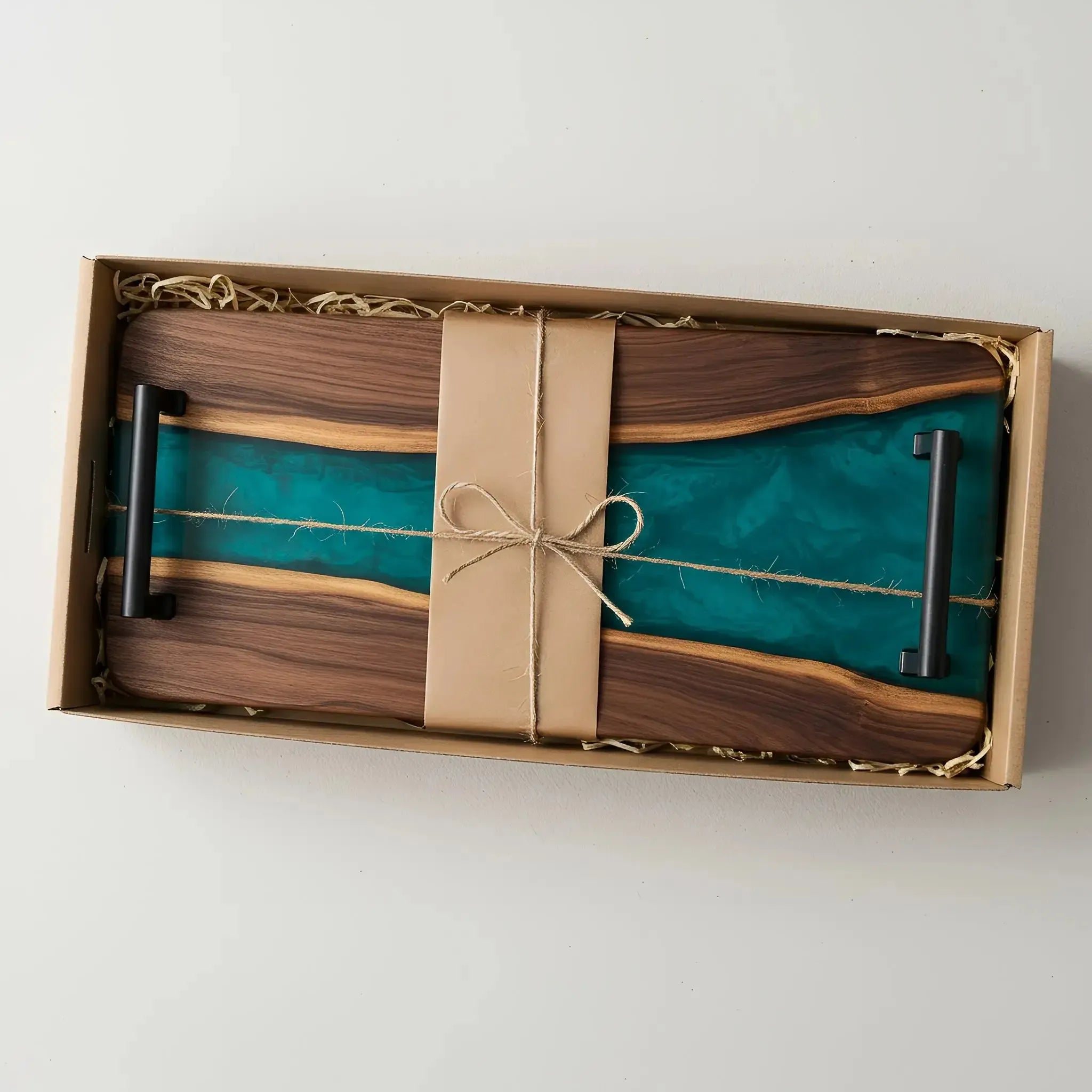 a charcuterie board carefully packaged for shipping. The board is made from live edge black walnut with a smooth, polished finish and a deep teal blue epoxy river running through the center. It’s wrapped in kraft paper and tied neatly with natural twine for a handmade, artisan feel. Matte black metal handles are attached at both ends.