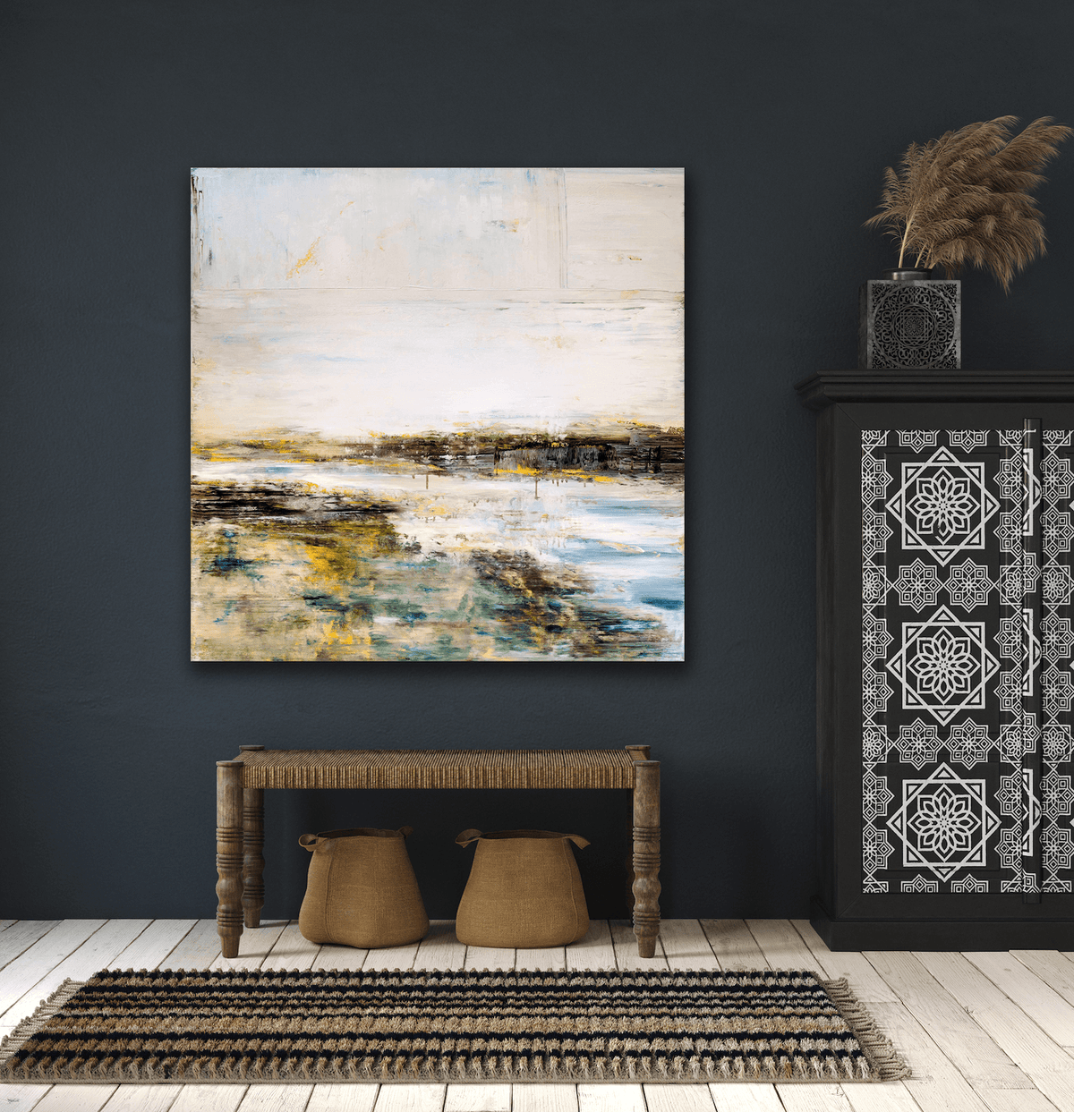 Abstract landscape canvas with shades of white, blue, brown, and yellow hangs on dark wall above rustic wooden bench and patterned rug.