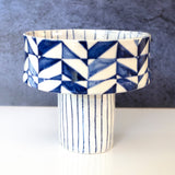 White ceramic serving bowl with a tall cylindrical base featuring vertical black stripes and a wide upper section decorated with blue geometric triangles forming a continuous pattern.