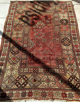 Rectangular antique Turkish wool prayer rug with a distressed red center panel featuring geometric and floral motifs, surrounded by intricate ivory, brown, and red patterned borders.
