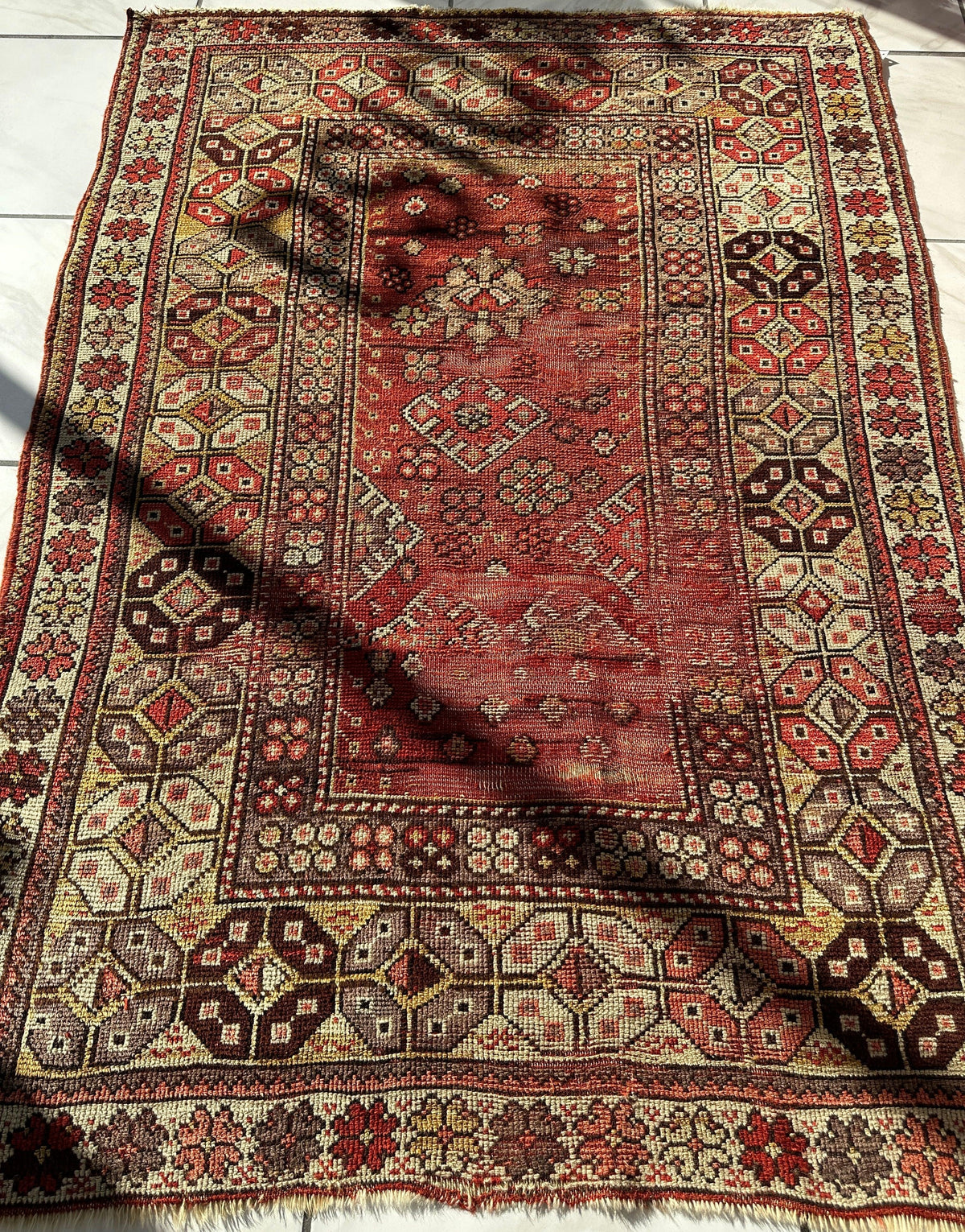 Rectangular antique Turkish wool prayer rug with a distressed red center panel featuring geometric and floral motifs, surrounded by intricate ivory, brown, and red patterned borders.