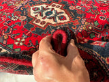 Hand lifting edge of a hand-knotted Persian Hamadan wool rug showing intricate traditional patterns in red, navy, cream, and green colors.