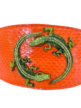 Close-up of an orange textured leather collar featuring two detailed brass alligator ornaments with green and red accents arranged in a circular pattern.