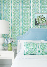 Bedroom corner with light blue upholstered headboard, patterned green and blue pillow matching vertical striped wallpaper, white side table with white lamp and small flower arrangement