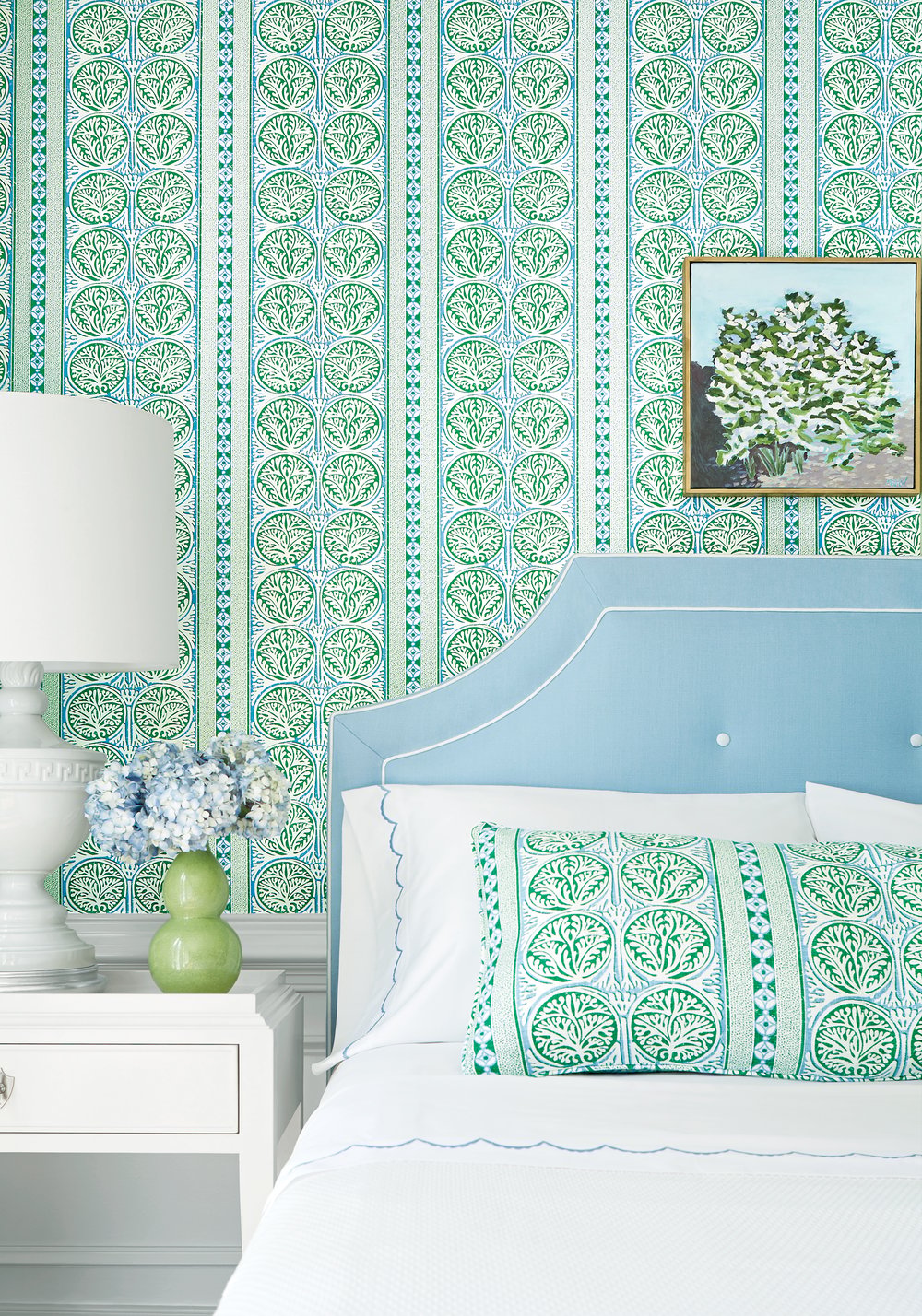 Bedroom corner with light blue upholstered headboard, patterned green and blue pillow matching vertical striped wallpaper, white side table with white lamp and small flower arrangement