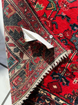 Close-up of a hand-knotted Persian Hamadan wool rug corner with intricate red, black, and cream geometric and floral patterns, showing a folded edge pinned to secure it.