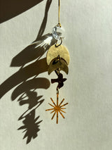 Hanging ornament with a clear vintage glass crystal, two overlapping brass crescent moons, a small bird silhouette, and a brass starburst pendant casting shadows on a light background.