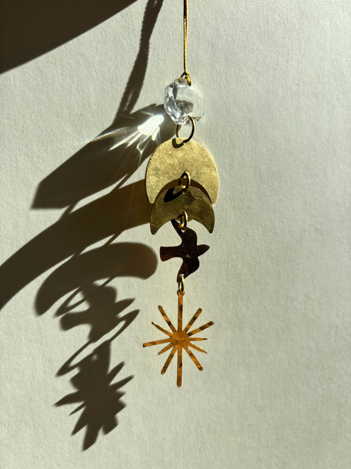 Hanging ornament with a clear vintage glass crystal, two overlapping brass crescent moons, a small bird silhouette, and a brass starburst pendant casting shadows on a light background.