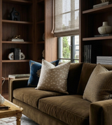 Brown velvet sofa with beige ivory pillow featuring a subtle feline pattern, flanked by textured gold and deep blue pillows in a warm wood-paneled room.