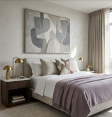 Bedroom with a beige upholstered bed, white and neutral-tone pillows including one with abstract geometric pattern, mauve throw, brass mushroom-shaped lamps on dark wood nightstands