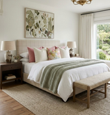Neutral bedroom with a beige grid-patterned headboard, white bedding accented by blush and floral pillows, soft sage green knitted throw, and a beige bench at bed's foot.