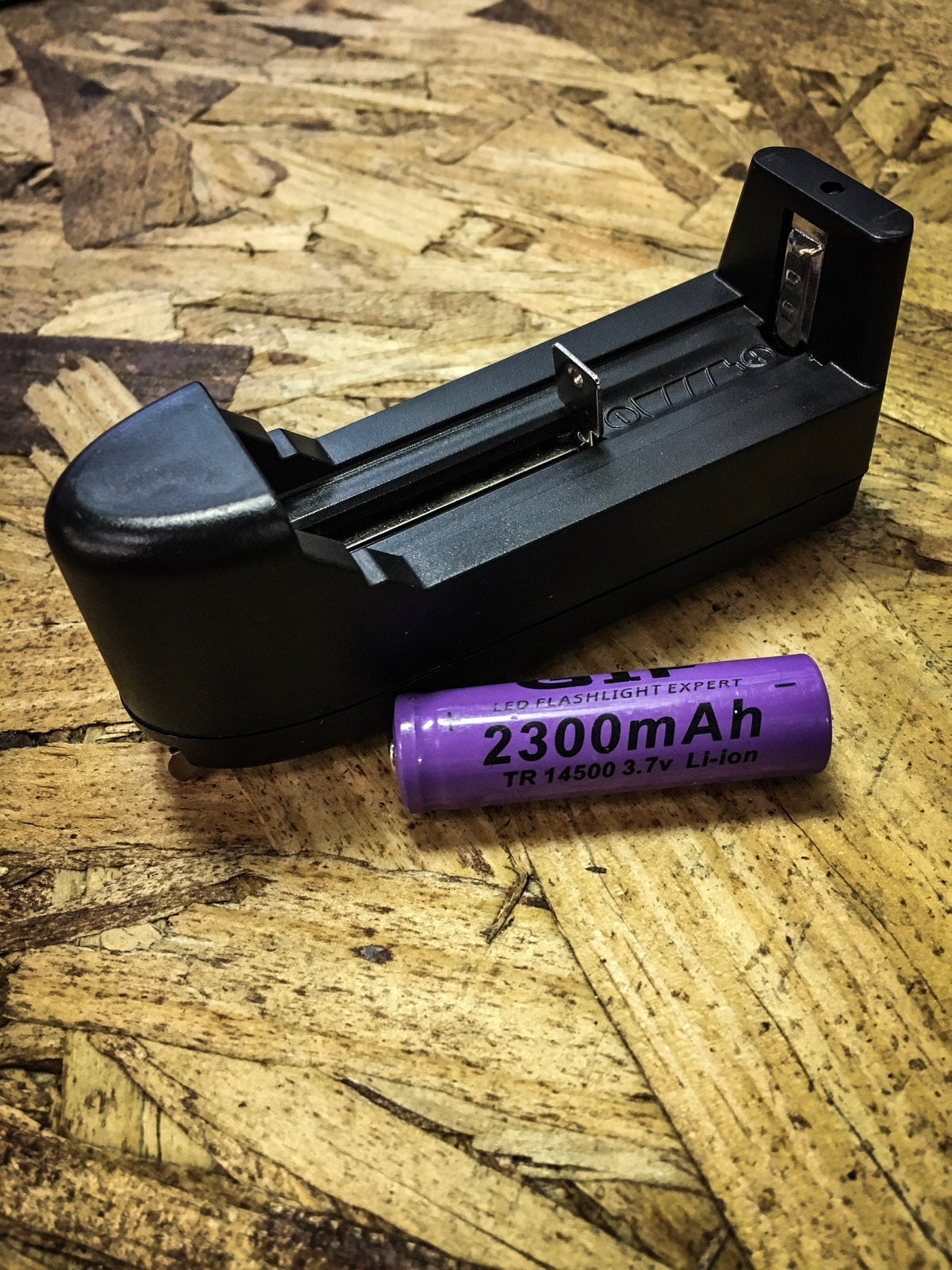 Black single-slot battery charger next to a purple 2300mAh TR 14500 3.7V Li-ion rechargeable battery on a wood-textured surface.