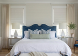 Bedroom with a blue upholstered headboard and white bedding accented by blue patterned and solid green pillows, flanked by white side tables with lamps and plants.