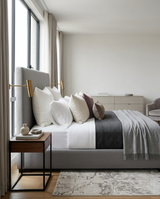 Neutral-toned bedroom with a light gray upholstered bed featuring multiple pillows in white, beige, and muted mauve, layered gray blankets, and a wooden side table with modern decor.