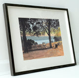 Framed watercolor painting of benches under leafy trees overlooking a calm river with a warm sunset sky in the background.