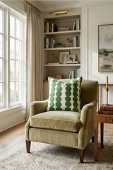 Light olive green armchair with tapered wooden legs, featuring a white pillow patterned with vertical rows of overlapping green circles, placed near a window and built-in shelves.