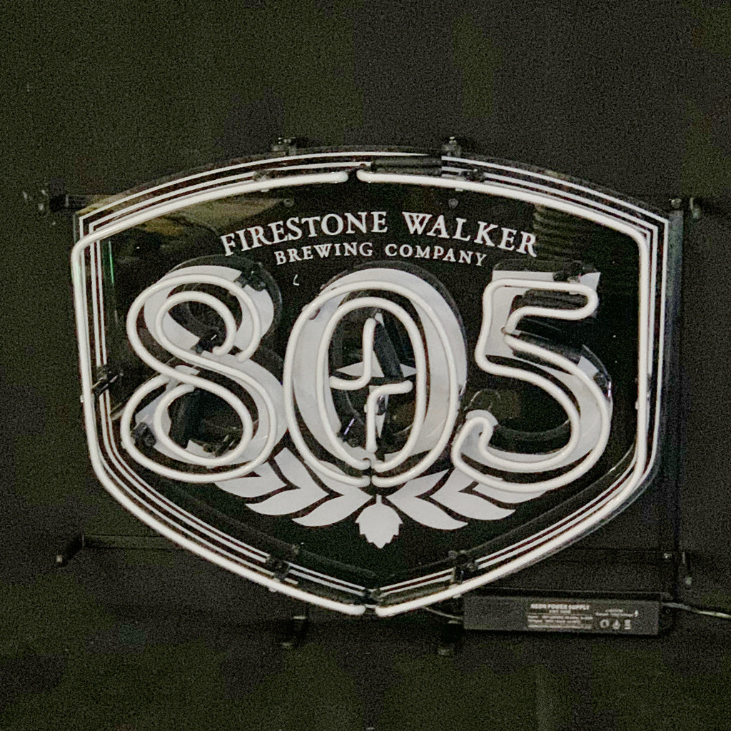 White neon sign shaped like a shield displaying 'Firestone Walker Brewing Company' above large numbers '805' with a hop flower design underneath on a dark background.