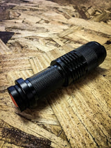 Compact black textured rechargeable flashlight with an orange power button, resting on a light brown wood particleboard surface.