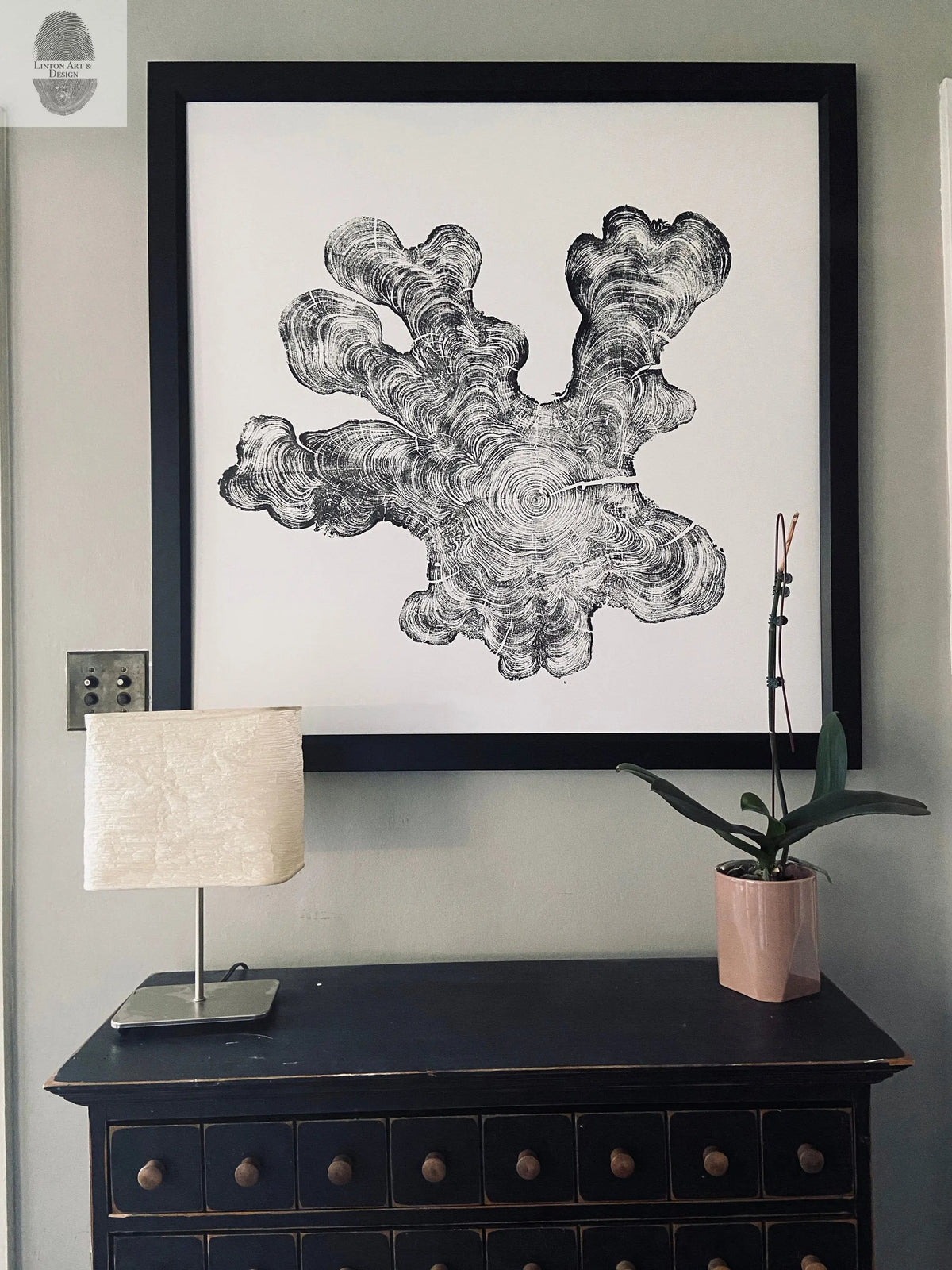 Black and white print of a detailed, irregularly shaped wood tree ring cross-section framed in black, hanging above a dark wooden cabinet with a lamp and potted orchid.