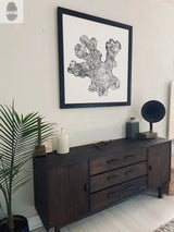 Black and white tree ring print with organic, irregular shape framed in black, hanging above dark wooden sideboard with decorative objects and a potted palm plant nearby.