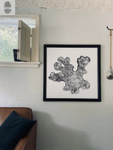 Black and white print of an irregularly shaped tree cross-section with detailed growth rings, framed in black and hung on a light gray textured wall above a brown leather couch.