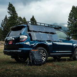 Dark blue SUV parked on grass with multiple Spectre 56W solar panels attached to its roof rack and side, a black backpack sits near the rear tire under cloudy sky.