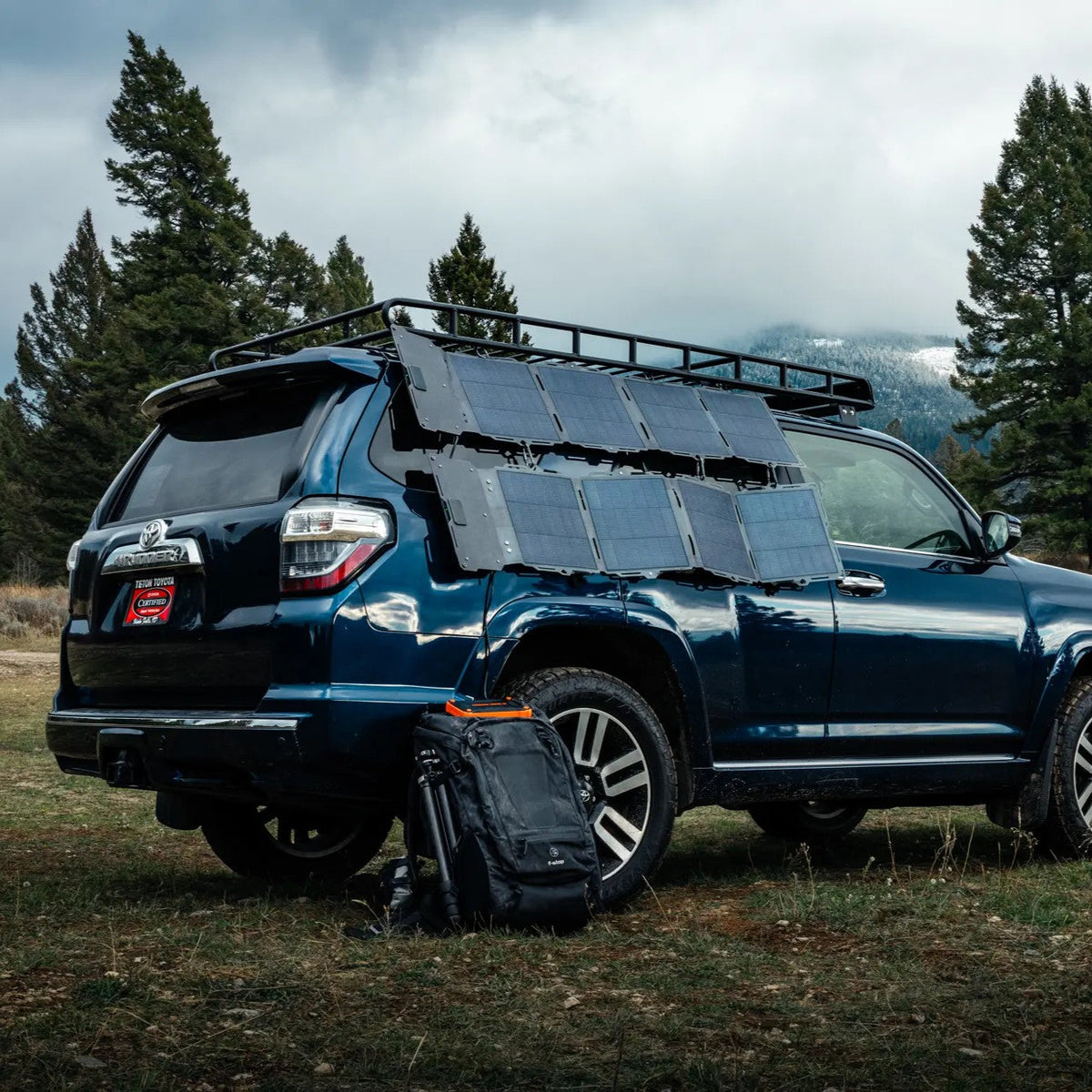 Dark blue SUV parked on grass with multiple Spectre 56W solar panels attached to its roof rack and side, a black backpack sits near the rear tire under cloudy sky.